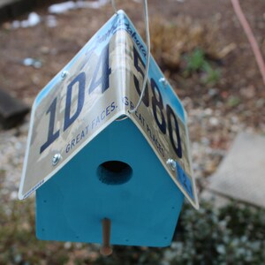 May include: A blue birdhouse made from a repurposed license plate from the state of Alaska. The license plate number is 1D4580 and has the text "Great Faces, Great Places".