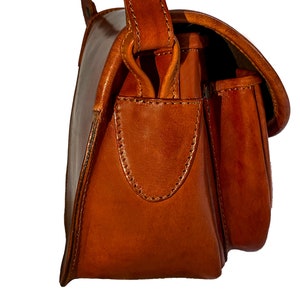 May include: A brown leather satchel bag with a flap closure and a small pocket on the front. The bag has a single strap and a buckle closure.