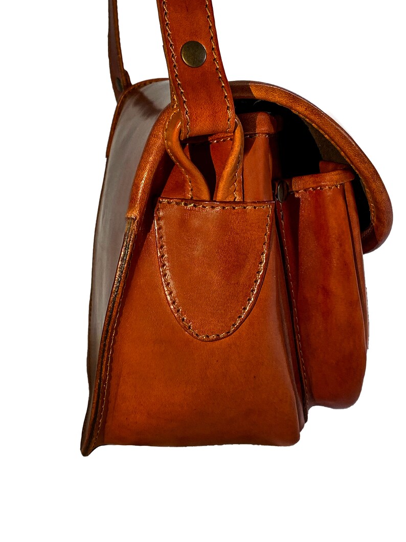 May include: A brown leather satchel bag with a flap closure and a small pocket on the front. The bag has a single strap and a buckle closure.