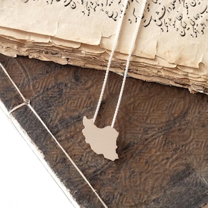 Puede incluir: Un collar con colgante plateado con un recorte en forma de país, sobre un libro antiguo con páginas envejecidas. El libro tiene una cubierta oscura y texturizada, y la cadena del collar es delicada.