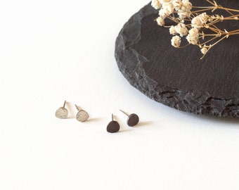 May include: A minimalist jewellery display featuring two pairs of stud earrings. One pair has a textured, half-moon design, and the other is a smooth, circular shape. The earrings are silver and dark grey. A black, round slate and dried flowers are in the background.