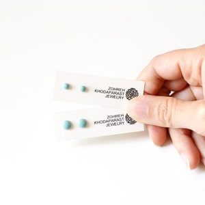 May include: Two cards with a pair of light blue round stud earrings on each. The cards have the text "Zohreh Khodaparast Jewelry" printed on them.