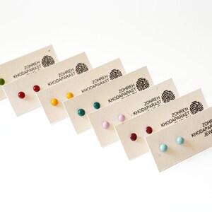 May include: Set of eight pairs of small, round stud earrings in various colors, including green, red, yellow, teal, pink, burgundy, light blue, and turquoise. The earrings are displayed on small cards with the text "Zohreh Khodaparast Jewelry".