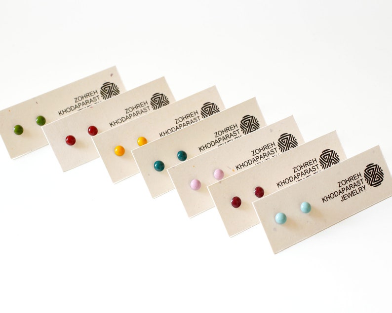 May include: Set of eight pairs of small, round stud earrings in various colors, including green, red, yellow, teal, pink, burgundy, light blue, and turquoise. The earrings are displayed on small cards with the text "Zohreh Khodaparast Jewelry".