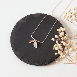 Puede incluir: Un collar de plata con un delicado colgante de diseño de hoja. El collar se muestra sobre una superficie de pizarra negra.