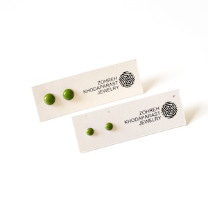 May include: Two pairs of olive green stud earrings on cream-coloured cards. The cards have the text "Zohreh Khodaparast Jewelry" and a black geometric logo. One pair is larger than the other.