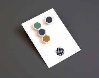 May include: Four hexagonal shaped earrings in shades of teal, blue, and gold. The earrings are displayed on a white card with a black geometric design.
