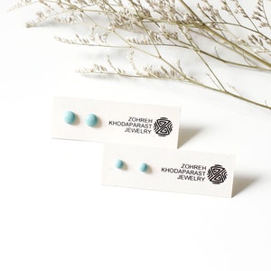 May include: A pair of light blue round stud earrings on a white card with the text "Zohreh Khodaparast Jewelry".