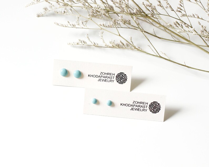 May include: A pair of light blue round stud earrings on a white card with the text "Zohreh Khodaparast Jewelry".