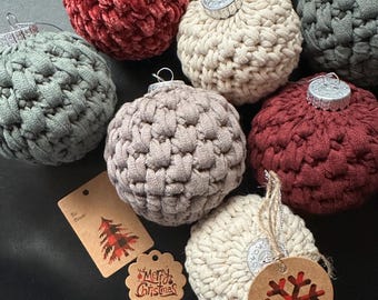 Op de afbeelding: Een verzameling handgemaakte kerstballen in verschillende kleuren, waaronder rood, crème en grijs. De ornamenten hebben een getextureerd, gehaakt uiterlijk en zijn afgewerkt met zilveren doppen. Sommige ballen hebben cadeaulabels met kerstthema's.