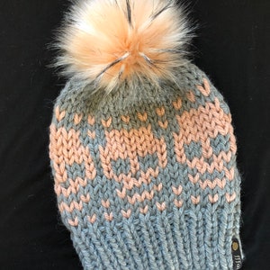 May include: A gray and pink knit beanie with a fluffy white and pink pom pom on top. The beanie has a small brown leather tag with the word "Fital" on it.