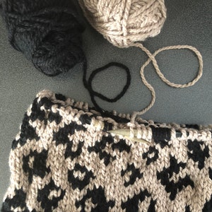 May include: A close-up of a knitting project in progress. The project is a black and beige patterned knit hat. Two balls of yarn, one black and one beige, are visible in the background.