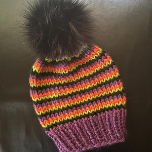 Könnte beinhalten: Eine gestrickte Mütze in Lila, Schwarz, Grün und Orange mit einem schwarzen Kunstfellbommel.