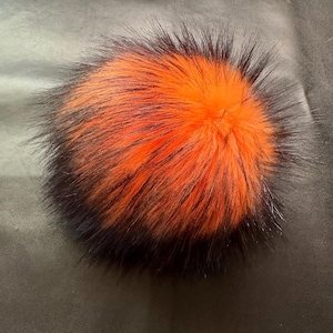 May include: A fluffy orange and black pom pom with long, soft fibres. The pom pom is round and has a gradient of colour from orange to black.
