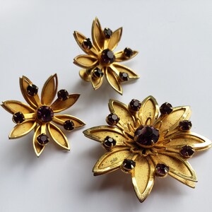 May include: A set of three gold-tone flower-shaped brooches with purple rhinestones.