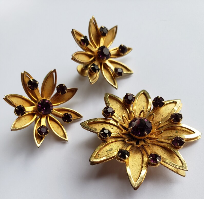 May include: A set of three gold-tone flower-shaped brooches with purple rhinestones.