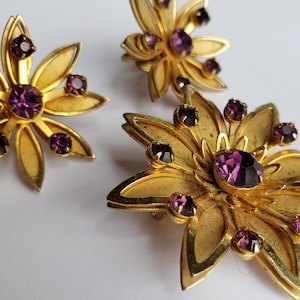 May include: A set of three gold-tone flower-shaped brooches with purple rhinestones.