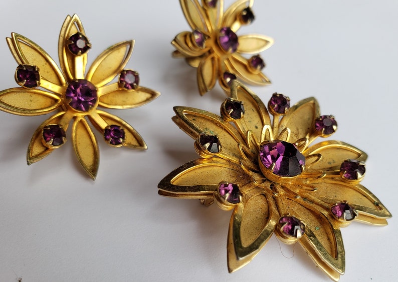 May include: A set of three gold-tone flower-shaped brooches with purple rhinestones.