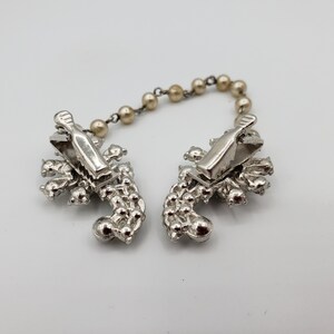 May include: A pair of silver-tone brooch clips connected by a beaded chain. Each clip features a cluster of rhinestones in a floral design.