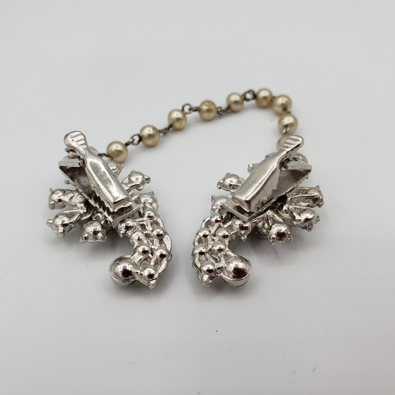 May include: A pair of silver-tone brooch clips connected by a beaded chain. Each clip features a cluster of rhinestones in a floral design.