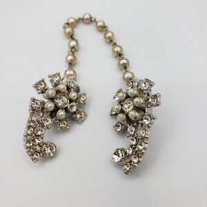 May include: A pair of silver tone jewellery clips with a pearl and rhinestone design. The clips are connected by a chain of faux pearls.