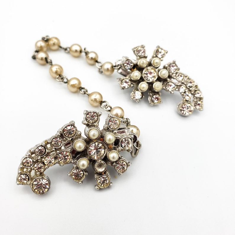 May include: A pair of vintage silver tone brooch clips with a pearl and rhinestone design. The clips are connected by a chain of faux pearls.