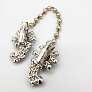 May include: A pair of silver tone scorpion shaped brooches with rhinestones connected by a chain of faux pearls.