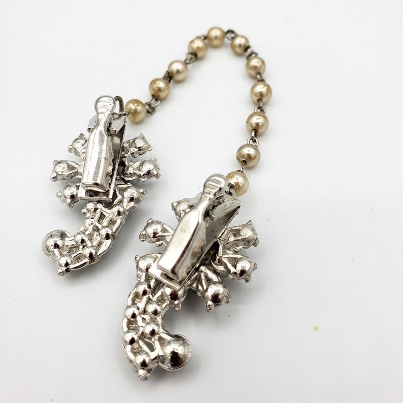 May include: A pair of silver tone scorpion shaped brooches with rhinestones connected by a chain of faux pearls.