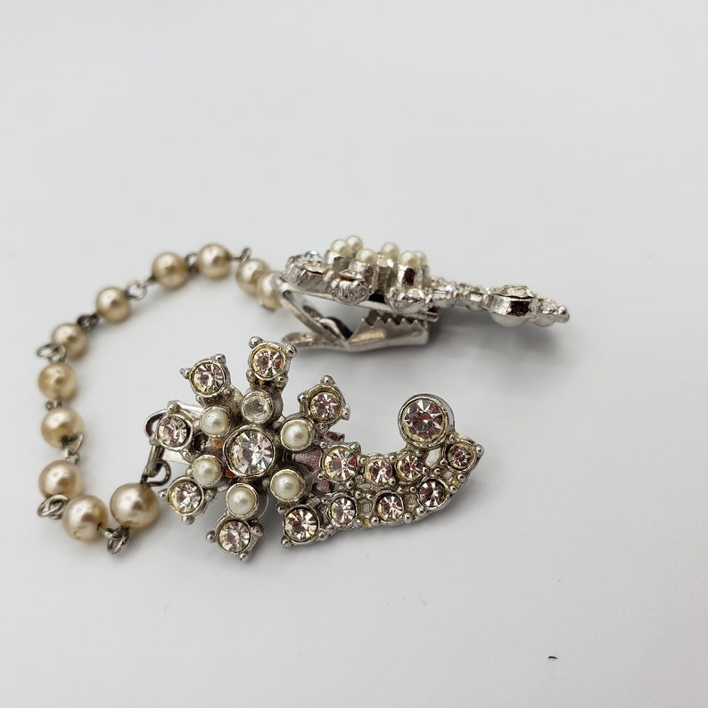 May include: A silver and crystal brooch with a pearl and rhinestone chain. The brooch features a floral design with a large crystal in the centre.