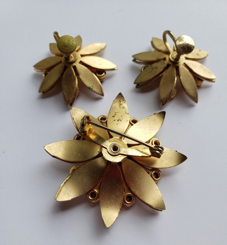 May include: A set of three gold-toned flower-shaped brooches. Two of the brooches are earrings with a post and back closure. The third brooch has a pin and clasp closure.