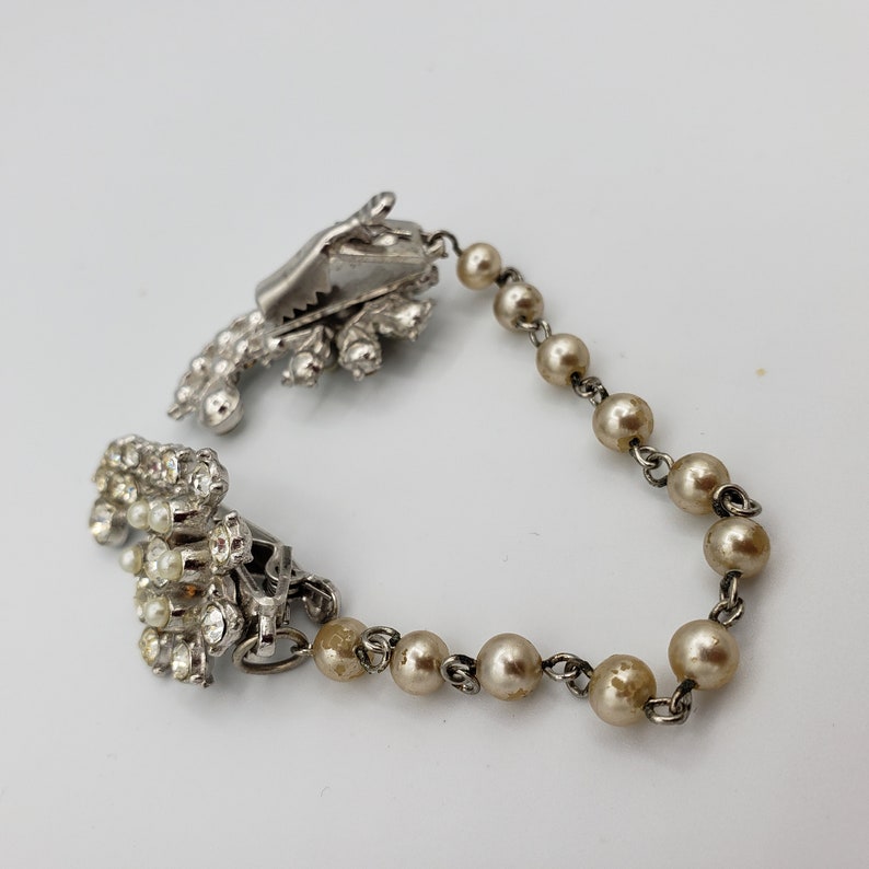 May include: A silver and pearl necklace with a rhinestone clasp. The necklace is made of small, round pearls and has a silver chain. The clasp is a cluster of rhinestones.
