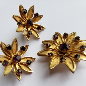May include: A set of three gold-tone brooches with purple rhinestones in the shape of flowers.