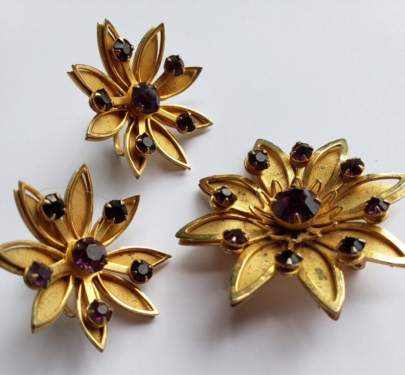 May include: A set of three gold-tone brooches with purple rhinestones in the shape of flowers.