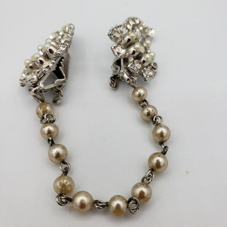 May include: A pair of silver tone, rhinestone and faux pearl brooches connected by a chain of faux pearls.