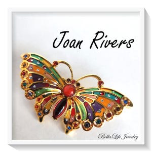 May include: A gold butterfly brooch with vibrant enamel wings and jewel detailing. The brooch displays the text "Joan Rivers" in black script. The brooch is set within a white frame.