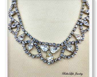 May include: A silver necklace with a large, teardrop-shaped rhinestone centerpiece surrounded by smaller rhinestones. The necklace has a delicate, feminine design.