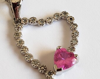 Peut inclure: Un collier avec un pendentif en forme de cœur argenté orné d'une pierre précieuse rose en forme de cœur au centre. Le pendentif est entouré de petites pierres précieuses claires.
