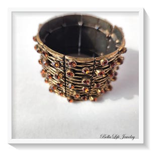 May include: A gold-toned stretch bracelet with a textured, woven design. The bracelet is adorned with numerous small, round, amber-colored gemstones. The Bella Life Jewelry logo is visible in the bottom right corner.