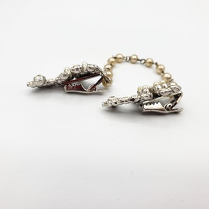 May include: A pair of silver tone clips with pearl accents connected by a chain of pearls. The clips are designed to hold fabric together.
