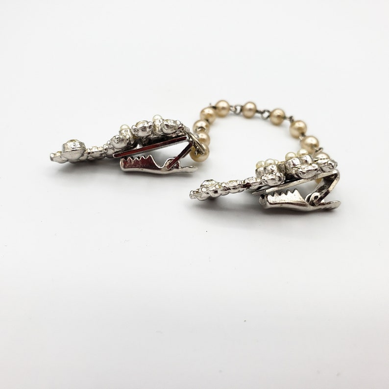 May include: A pair of silver tone clips with pearl accents connected by a chain of pearls. The clips are designed to hold fabric together.