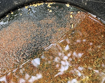 May include: A black gold pan filled with water and sediment. The sediment is a mix of brown, tan, and yellow, with small flecks of gold visible. The pan's rim is visible at the top, and the water reflects light.