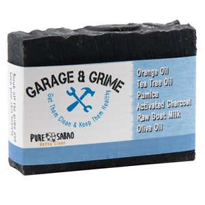 May include: A dark gray bar of soap with the text "GARAGE & GRIME" and a blue wrench and hammer logo. The label lists ingredients like orange oil and activated charcoal. The soap is designed to scrub off grime.