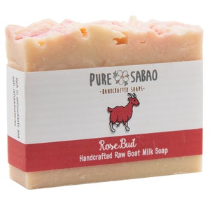 May include: A bar of pink and white goat milk soap with a label that reads "PURE SABAO - HANDCRAFTED SOAPS - Rose Bud Handcrafted Raw Goat Milk Soap".