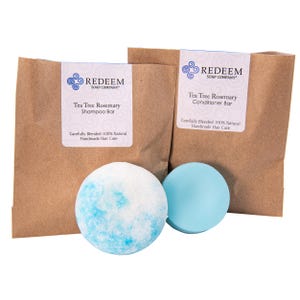 May include: Two brown paper bags with labels reading "Tea Tree Rosemary Shampoo Bar" and "Tea Tree Rosemary Conditioner Bar." A blue and white shampoo bar and a solid blue conditioner bar are in front of the bags. The labels state "Carefully Blended 100% Natural Handmade Hair Care."