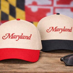 May include: Two baseball caps with "Maryland" embroidered in red script. One cap has a red bill, the other a black bill. The caps are cream-colored with a red or black bill. Sunglasses rest on a wooden surface.