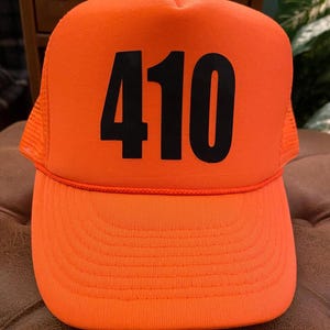 May include: Bright orange trucker hat with the number "410" printed in bold black letters across the front. The hat features a mesh back and a curved brim. A casual accessory.