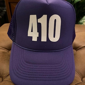 May include: A purple trucker hat with the number "410" in white block letters on the front. The hat has a mesh back and a curved brim. The hat is sitting on a brown surface.
