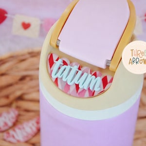 Puede incluir: Un vaso morado claro con tapa rosa claro y asa amarilla. El nombre "tatum" est&aacute; escrito en escritura azul claro. El vaso tiene un patr&oacute;n decorativo rojo y blanco. Una pancarta con corazones est&aacute; en el fondo.
