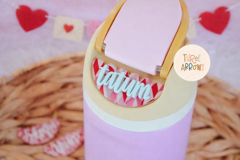 Puede incluir: Un vaso morado claro con tapa rosa claro y asa amarilla. El nombre "tatum" est&aacute; escrito en escritura azul claro. El vaso tiene un patr&oacute;n decorativo rojo y blanco. Una pancarta con corazones est&aacute; en el fondo.
