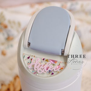 May include: A white water bottle with a light blue lid and a clear sticker with the name "Alycee" in pink lettering and colorful glitter. The sticker is on the front of the bottle, and the text "THREE Arrows COLLECTIVE" is on the side of the bottle.